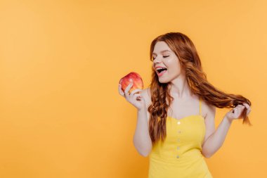 beautiful redhead girl biting apple isolated on yellow with copy space clipart