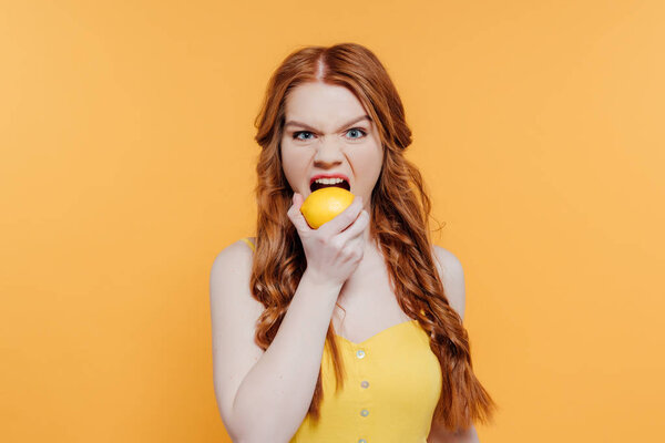 redhead girl looking at camera, making facial expression and biting lemon isolated on yellow
