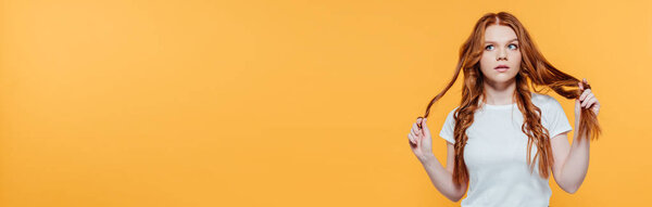 panoramic shot of beautiful redhead girl touching hair and looking away isolated on yellow with copy space