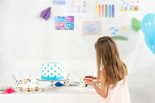 back view of adorable kid holding plate with donuts at birthday table with cake during party