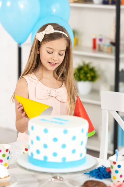adorable kid holding party cap near table with cake during birthday celebration