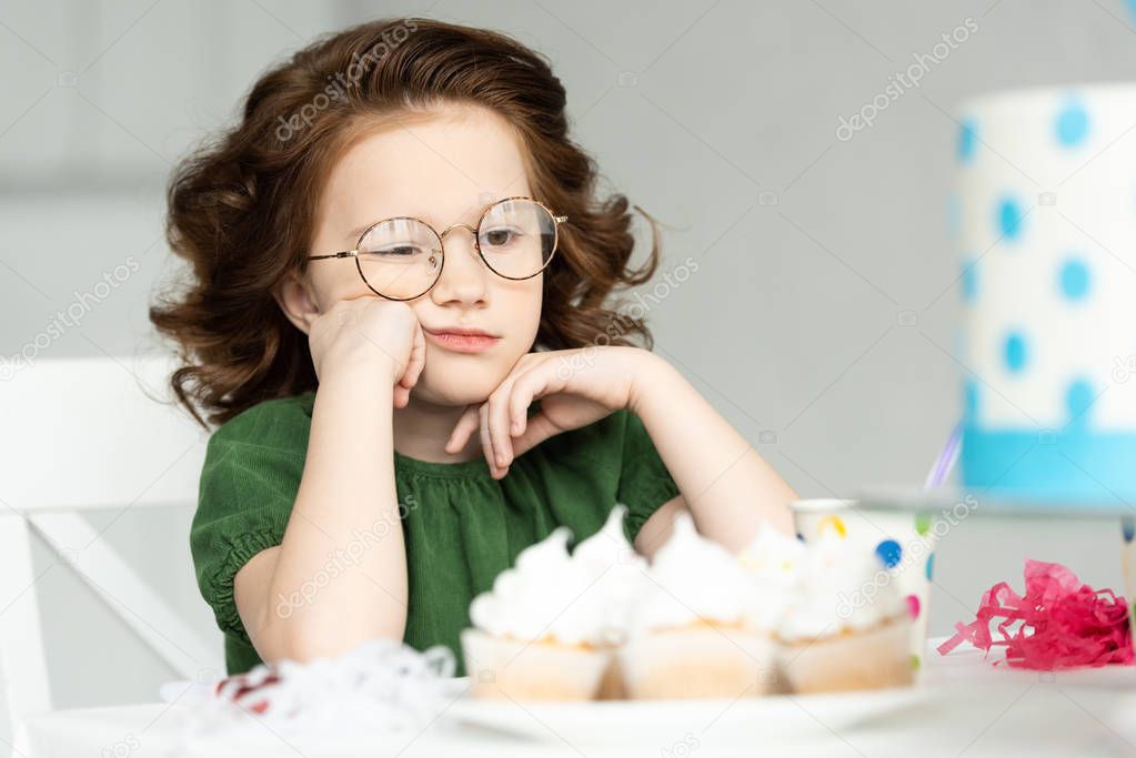 adorable bored kid propping chin while sitting at table with cupcakes
