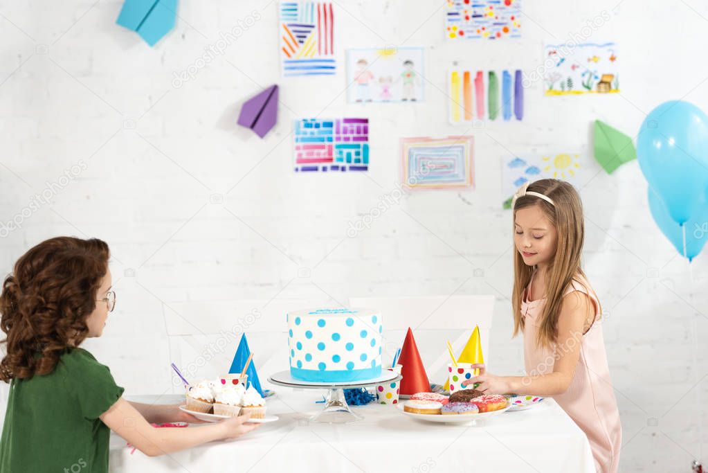 adorable kids sitting at party table with cake and cupcakes during birthday celebration