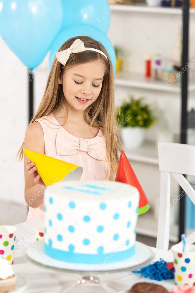 adorable kid holding party cap near table with cake during birthday celebration