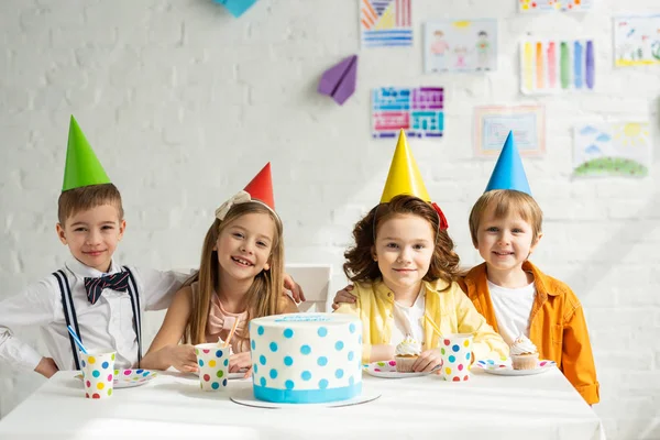 happy kids in party hats sitting at table with cake and looking at camera while celebrating birthday together