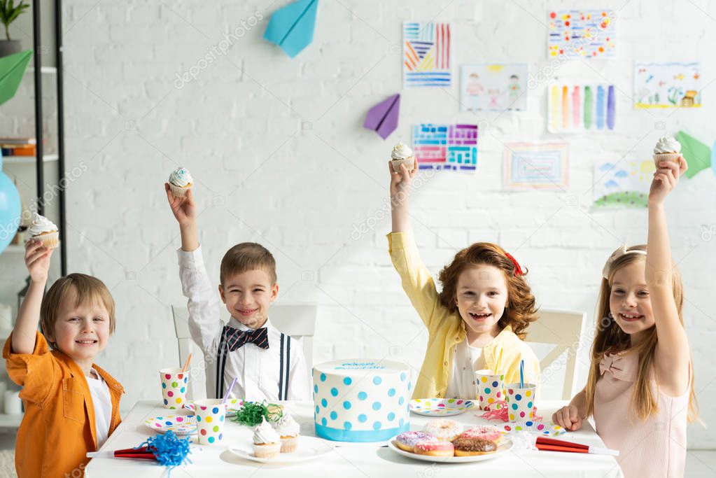 adorable kids sitting at table and holding cupcakes while celebrating birthday together