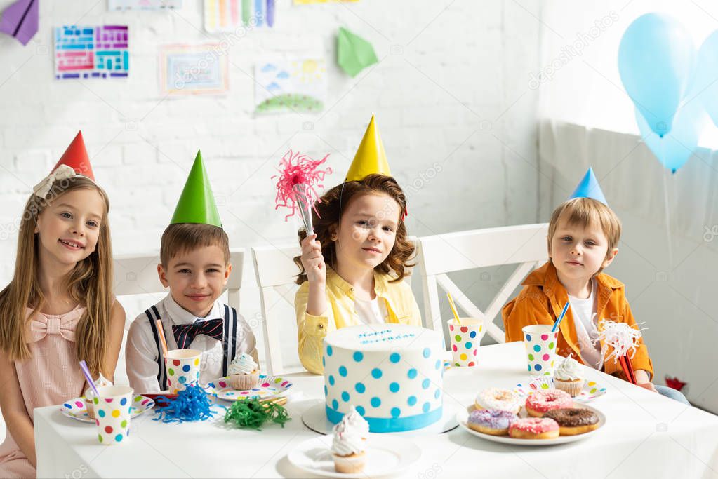 kids in party hats sitting at table and looking at camera during birthday celebration