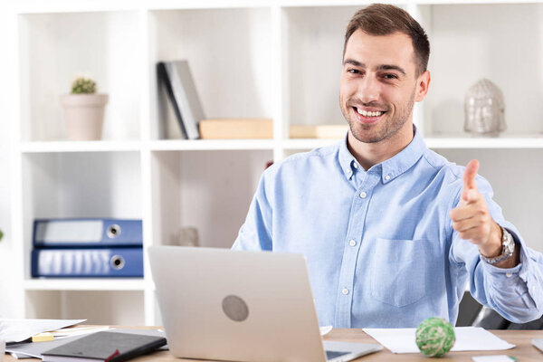 веселый бизнесмен показывает пальцем и смотрит в камеру рядом с ноутбуком
 
