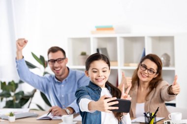 ofiste gesturing mutlu ebeveynlerin yanında akıllı telefonda selfie çeken neşeli çocuğun seçici odak 