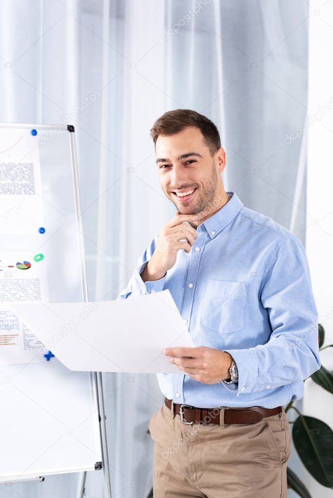 cheerful businessman standing near white board and holding blank paper 