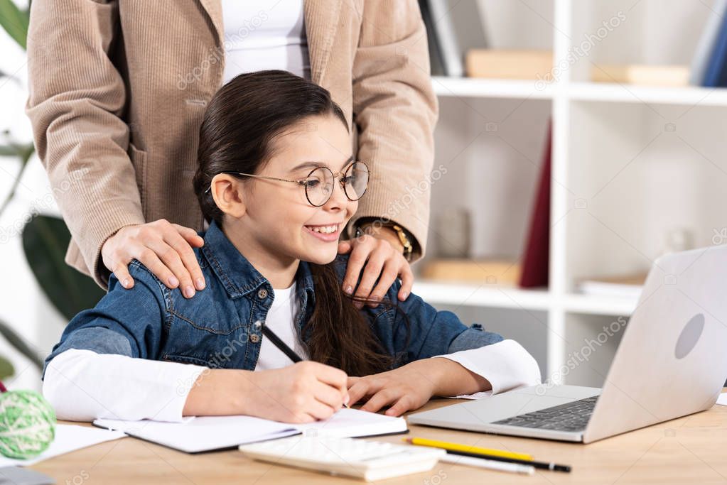 cropped view of mother putting hands on shoulders of happy kid looking at laptop in office 