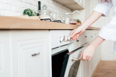 partial view of young woman in white shirt opening oven in kitchen clipart