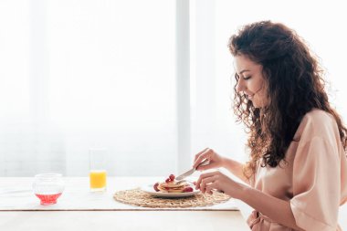 side view of smiling girl eating pancakes in kitchen clipart