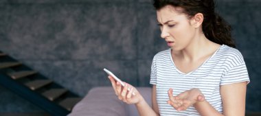 panoramic shot of irritated woman holding smartphone at home clipart