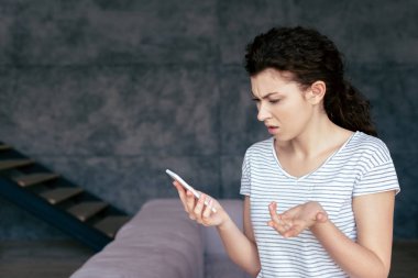irritated woman in t-shirt using smartphone in living room clipart