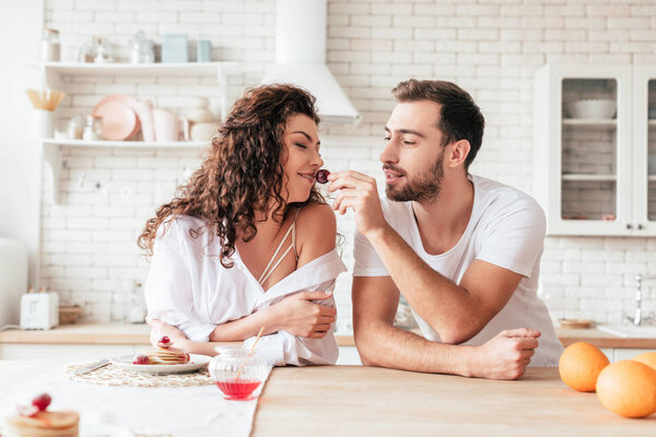 bearded handsome man feeding girlfriend with berry in kitchen
