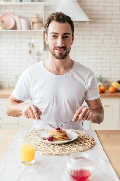 キッチンでパンケーキを食べる白いTシャツで笑顔のあごひげの男 — ストック写真