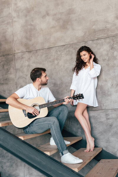 smiling bearded man sitting on stairs and playing acoustic guitar to girlfriend