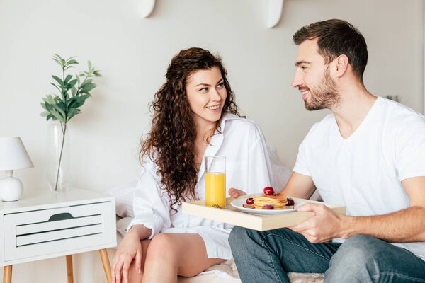 smiling couple with tray with breakfast looking at each other in bedroom