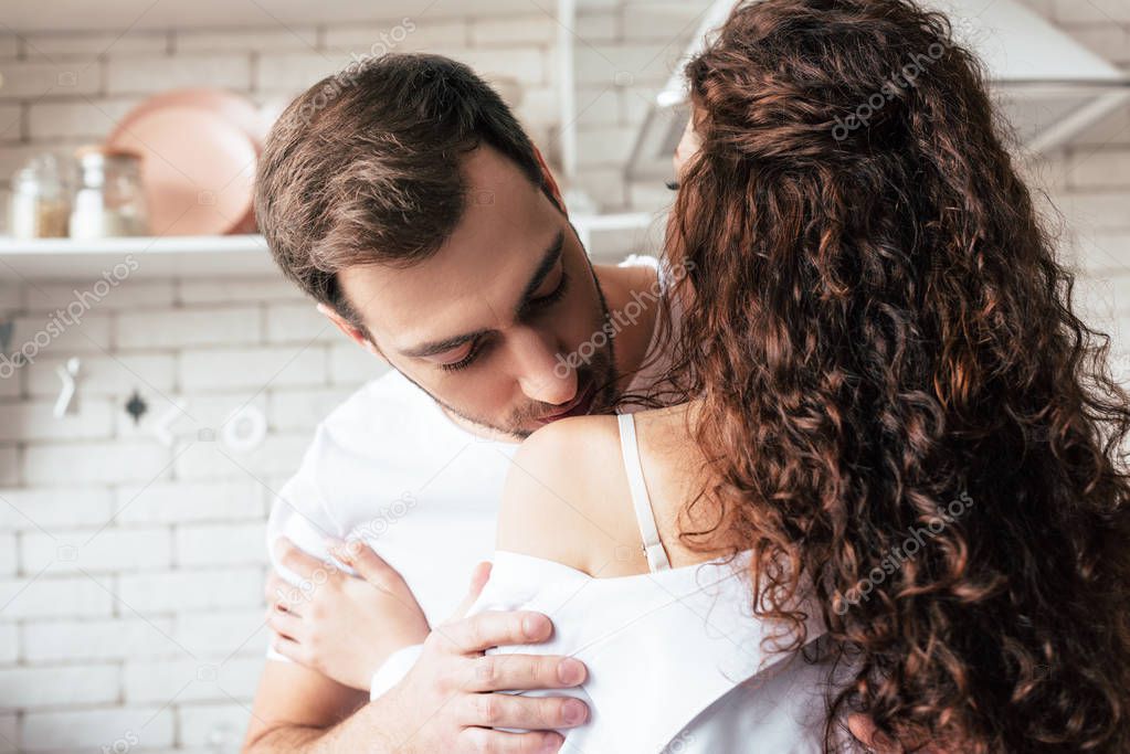 man kissing curly girlfriend in shoulder in kitchen