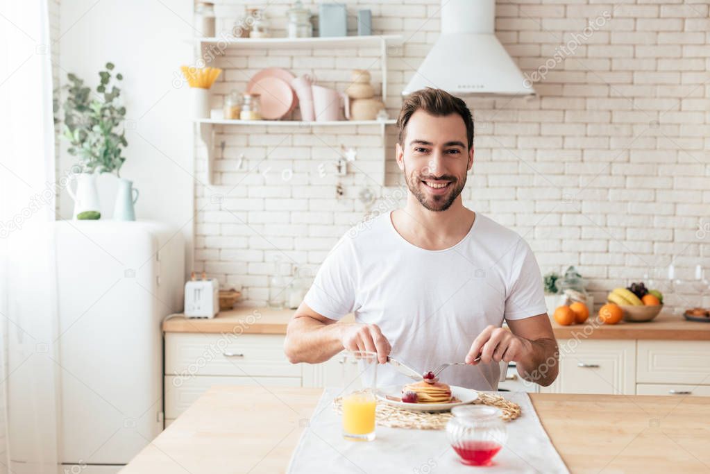 smiling bearded man in white t-shirt eating pancakes in kitchen