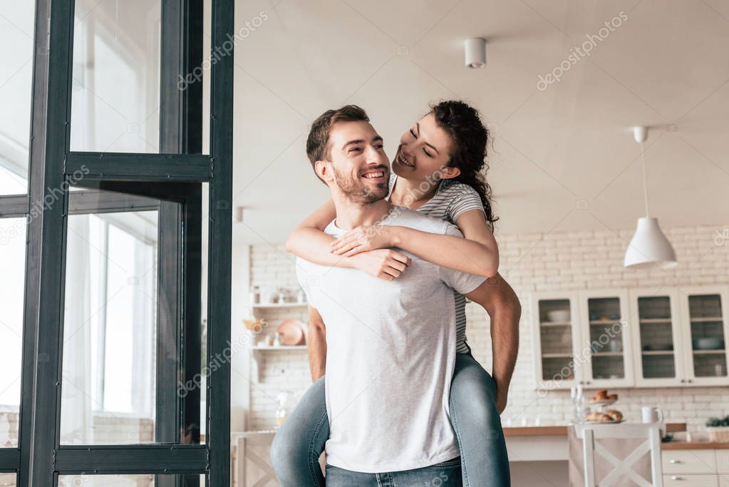 laughing bearded man carrying girlfriend piggyback at home