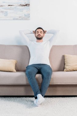 handsome bearded man with eyes closed and Hands Behind Back resting on couch at home clipart