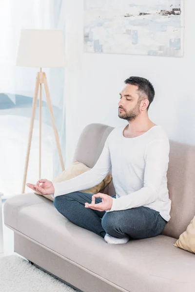handsome man with eyes closed sitting on couch in Lotus Pose and meditating at home