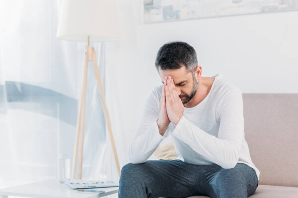 handsome bearded man with eyes closed doing please gesture and praying at home