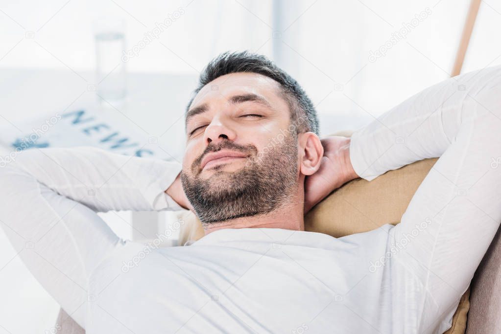 selective focus of handsome bearded man with eyes closed and Hands Behind Back resting on couch at home