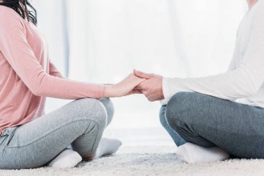 cropped view of man and woman in casual clothes sitting in lotus pose, meditating and holding hands at home clipart