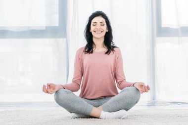 beautiful smiling woman in Lotus Pose practicing meditation in Living Room at home  clipart