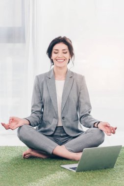 smiling businesswoman in suit meditating while sitting on grass mat in Lotus Pose near laptop 
