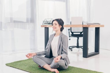 takım elbise güzel mutlu iş kadını Lotus Pose çim mat üzerinde oturan ve ofiste meditasyon