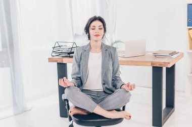 beautiful businesswoman in suit sitting on chair and meditating in Lotus Pose in office clipart