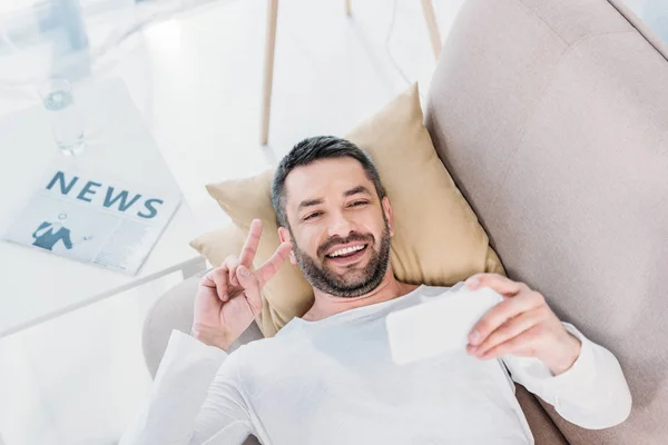 ソファに横たわり 自宅でスマートフォンで自分撮りを取るハンサムな笑顔の男 — ストック写真