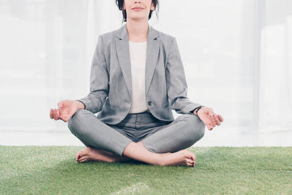 Cropped view of businesswoman in suit meditating while sitting on grass mat in Lotus Pose 