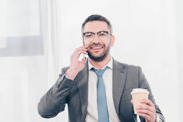 handsome smiling businessman in glasses and suit with coffee to go talking on smartphone