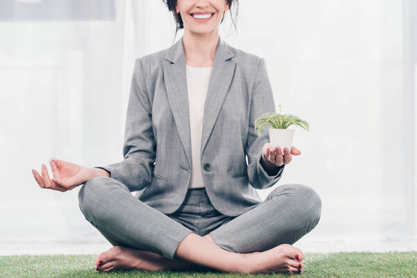 cropped view of smiling businesswoman in suit meditating on grass mat in Lotus Pose with flowerpot 