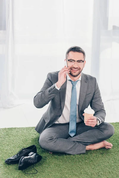 英俊的微笑商人在西装坐在草垫上 在智能手机上交谈 拿着咖啡去 — 图库照片