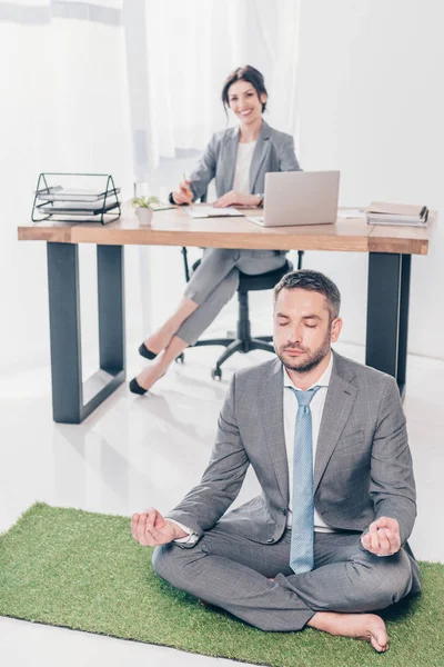 Businessman Meditating Lotus Pose Grass Mat While Businesswoman Sitting Table — Stock Photo, Image