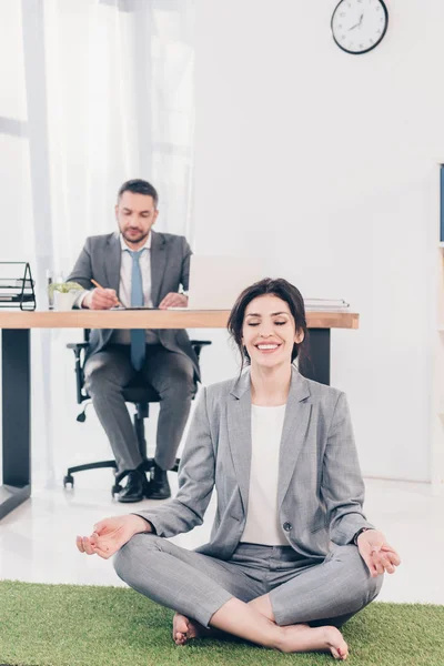 ビジネスマンがテーブルに座り オフィスの机に書いている間 草のマットの上でロータスポーズで瞑想する笑顔のビジネスウーマン — ストック写真