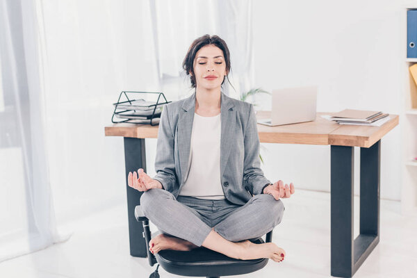 красивая деловая женщина в костюме сидит на стуле и медитирует в Lotus Pose в офисе
