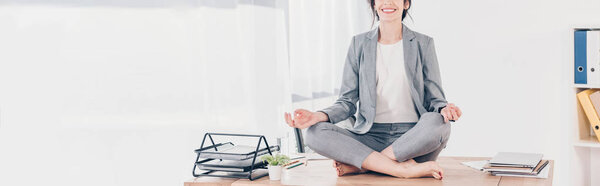 панорамный снимок деловой женщины в костюме, сидящей на столе и медитирующей в Lotus Pose в офисе
