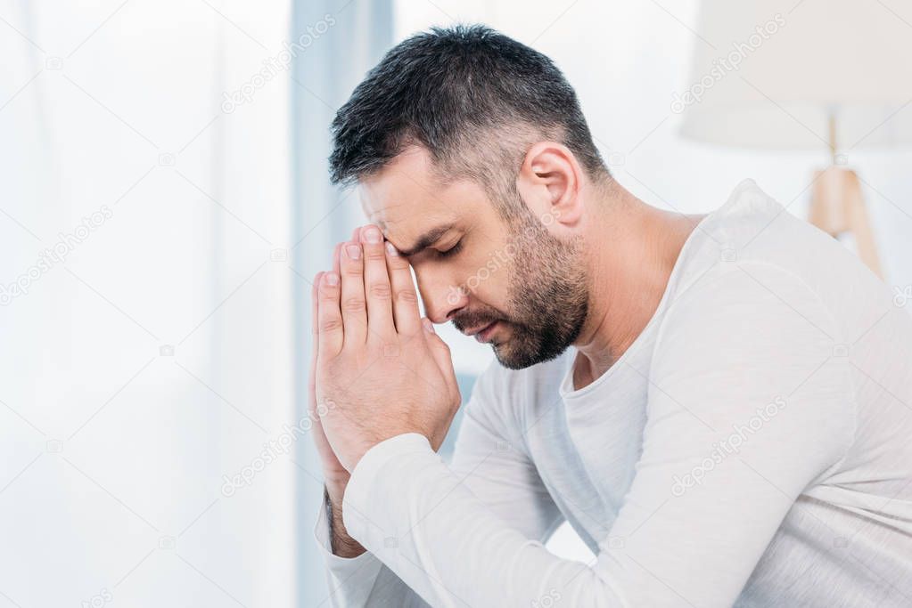 handsome bearded man with eyes closed doing please gesture and praying at home