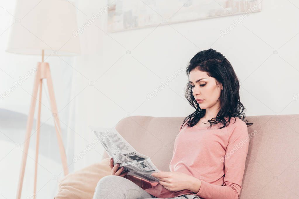 beautiful serious woman sitting on couch and reading newspaper at home