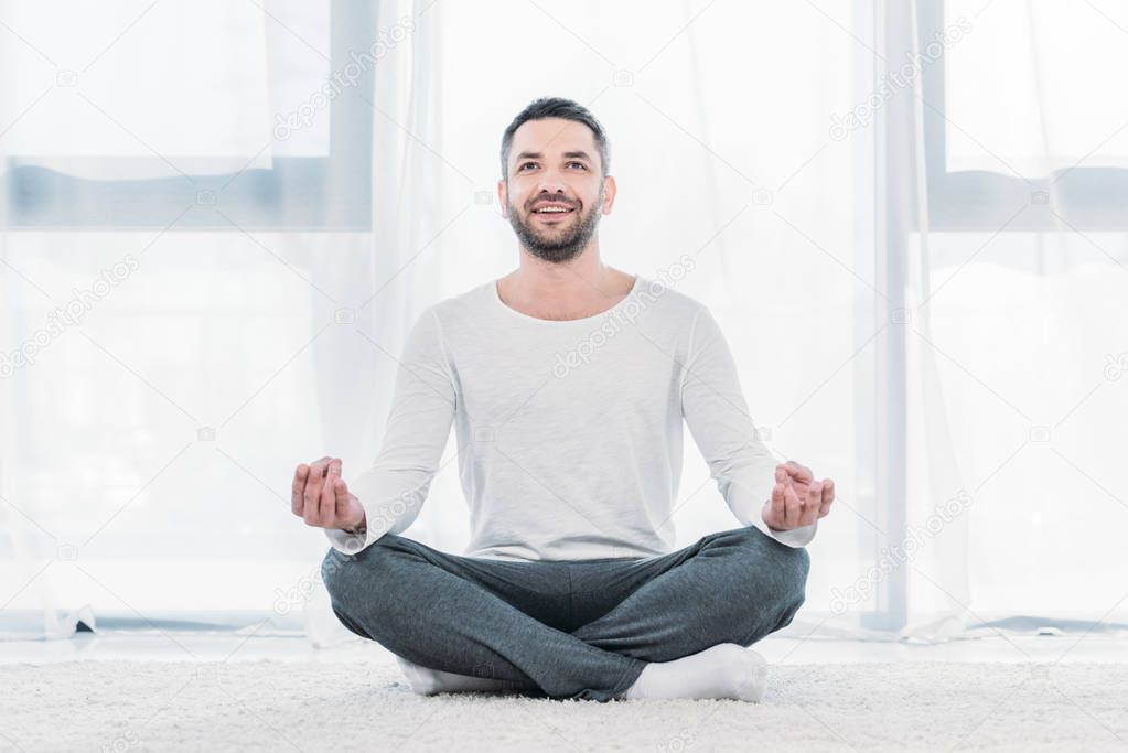 happy handsome man sitting on carpet in Lotus Pose and meditating at home