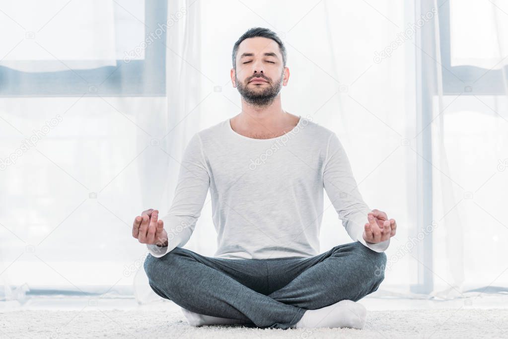 good-looking man with eyes closed sitting on carpet in Lotus Pose and meditating at home