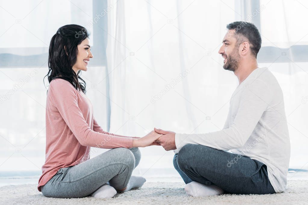 smiling man and woman in casual clothes sitting in lotus pose, meditating and holding hands at home