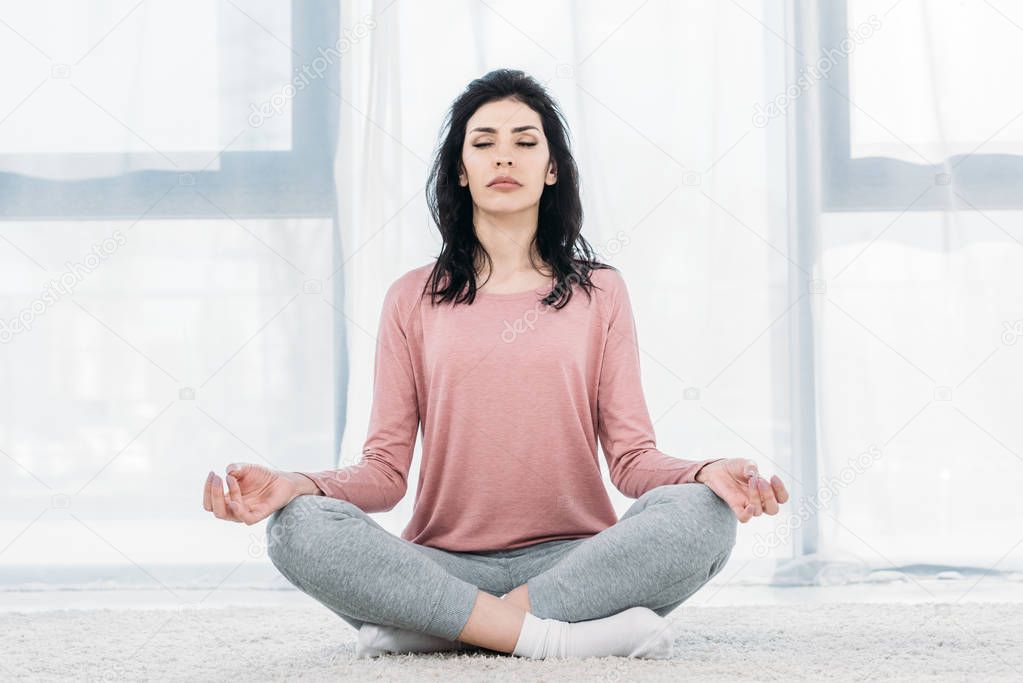 beautiful woman with eyes closed in Lotus Pose practicing meditation in Living Room at home 